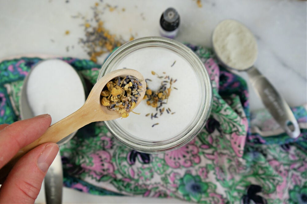 Wooden spoon pouring chamomile and lavender into a jar with salt.