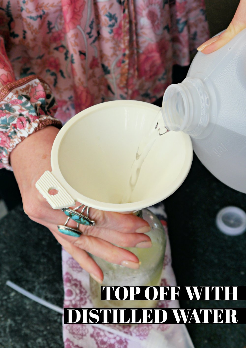 Hand pouring a jug of distilled water into a funnel over a glass spray bottle.